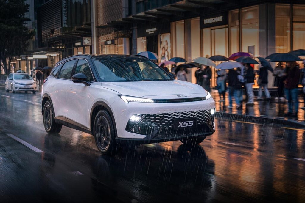 Front three quarter view of a white BAIC X55 SUV driving through a busy city street at night in the rain